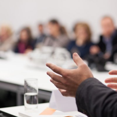 A business-man's hands gesture. In the background are people gathered around a conference 